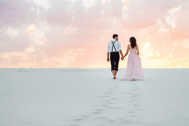 Jong stel een man in zwarte broek en een meisje in een roze jurk lopen langs het witte zand van de woestijn