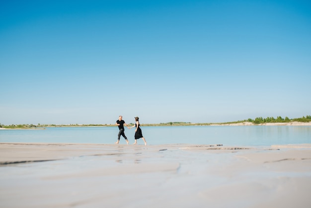 Jong stel een man met een meisje in zwarte kleren loopt op het witte zand aan de rand van blauw water