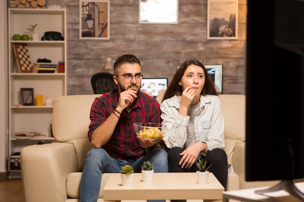Jong stel kijkt geconcentreerd op tv terwijl ze een film kijken en chips eten. Paar zittend op de bank.