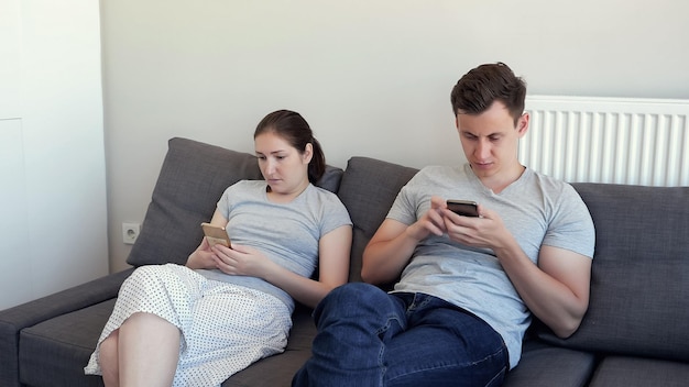 Jong stel, man en vrouw kijken in hun mobiele telefoons zittend op de bank in hun woonkamer. Negeer elkaar. Smartphone vervangt live communicatie, gadgetverslaving.