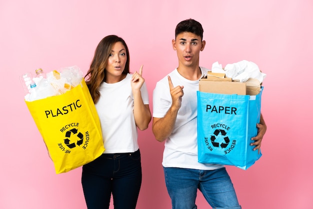 Jong stel met een zak vol plastic en papier op roze met de bedoeling de oplossing te realiseren terwijl ze een vinger opheffen