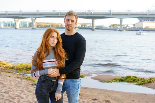 Jong stel van man en vrouw met lang rood haar van blanke etniciteit in vrijetijdskleding staan op het strand