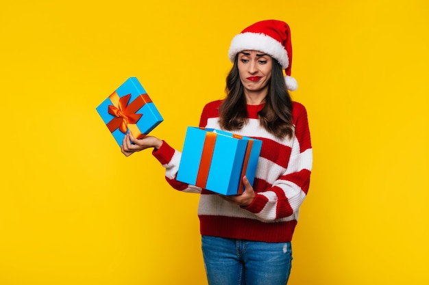 Jong teleurgesteld schattig meisje in kerstmuts pakt geschenkdoos uit op gele achtergrond met ongelukkig gezicht