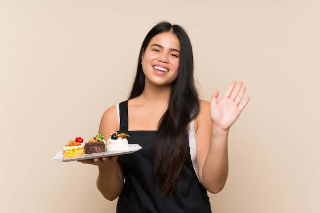 Jong tiener Aziatisch meisje die veel verschillende minicakes over geïsoleerde muur groeten met hand met gelukkige uitdrukking