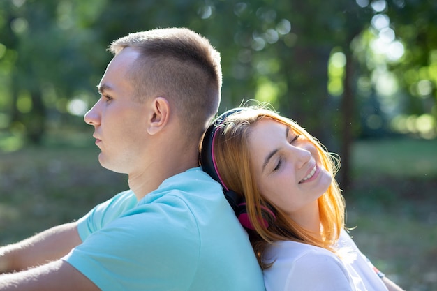 Jong tienerpaar samen in openlucht in de zomerpark. Meisje met rood haar luisteren naar muziek in oortelefoons en jongen verveelt zich.