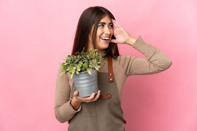 Jong tuinmanmeisje dat een installatie houdt die op roze muur wordt geïsoleerd die veel glimlacht