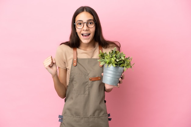 Jong tuinmanmeisje dat een plant houdt die op roze achtergrond wordt geïsoleerd die een overwinning in winnaarspositie viert