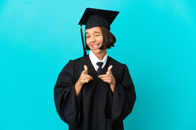 Jong universitair gediplomeerd meisje over geïsoleerde blauwe achtergrond die naar voren wijst en glimlacht