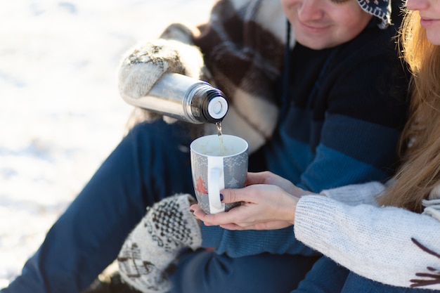 Jong verliefd paar drinkt een warm drankje uit een thermosfles, zit in de winter in het bos, verstopt in warme, comfortabele tapijten en geniet van de natuur. de man giet een drankje uit een thermoskan in een kopje