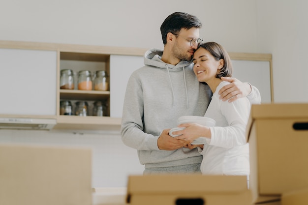 Jong verliefd paar omhelst en kust met zachte, houdt stapel witte borden vast, staat tijdens verhuisdag in de keuken, omringd met veel kartonnen dozen gevuld met persoonlijke bezittingen, spullen uitpakken