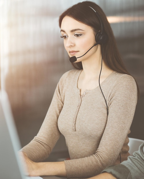 Jong vriendelijk meisje in headsets praat met de klant van een bedrijf, zittend aan de balie in een zonnig kantoor. Callcentermedewerkers aan het werk