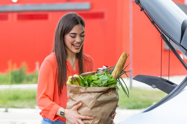 Jong vrouwen berijdend boodschappenwagentjehoogtepunt van voedsel op het openluchtparkeren. Jonge vrouw in parkeergarage, die het winkelen in laars van auto laden.