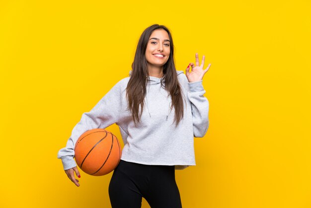 Jong vrouwen speelbasketbal over geïsoleerde gele muur die ok teken met vingers tonen