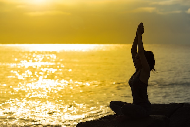 Jong vrouwensilhouet die en yoga op het strand bij zonsondergang mediteren uitoefenen.