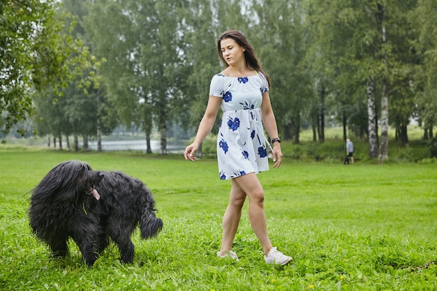 Jong wit meisje in jurk loopt overdag met zwarte ruige hond in openbaar park.