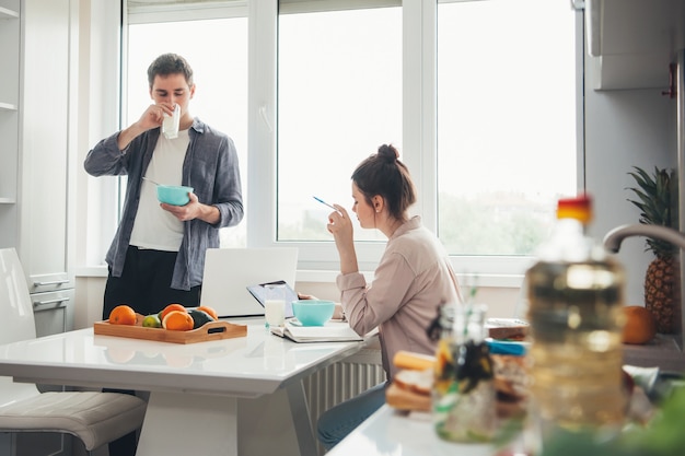 Jong zakenpaar in de keuken drinken en eten tijdens het werken aan elektronische apparaten