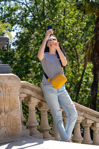 Foto jonge 30 jaar oude vrouw in jeans tshirt en gele tas wandelen in een park ciutadella in barcelona en het nemen van foto op haar smartphone reizen in spanje