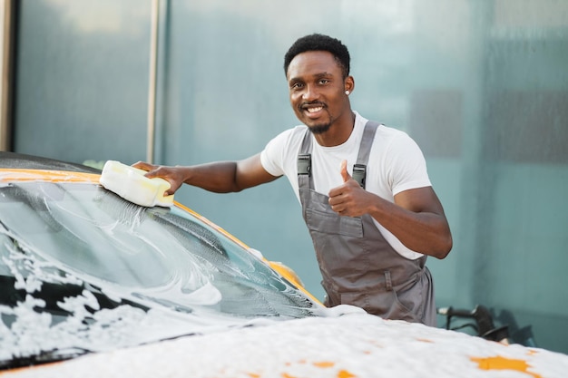 Jonge aantrekkelijke Afro-Amerikaanse man die zijn auto wast met schoonmaakschuim en gele spons