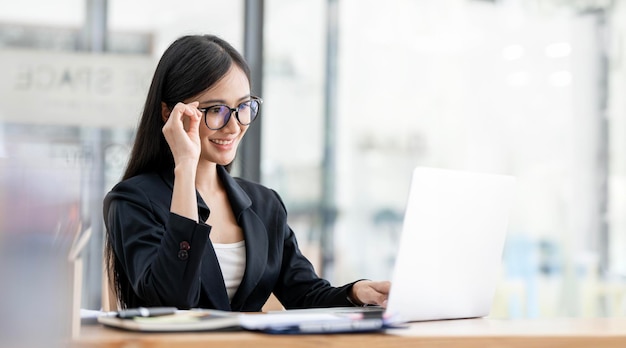 Jonge aantrekkelijke Aziatische onderneemster die met glazen aan laptop computer bij haar bureau werken