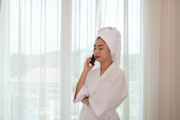 Jonge aantrekkelijke Aziatische vrouw in een badjas die in de woonkamer aan de telefoon staat en praat