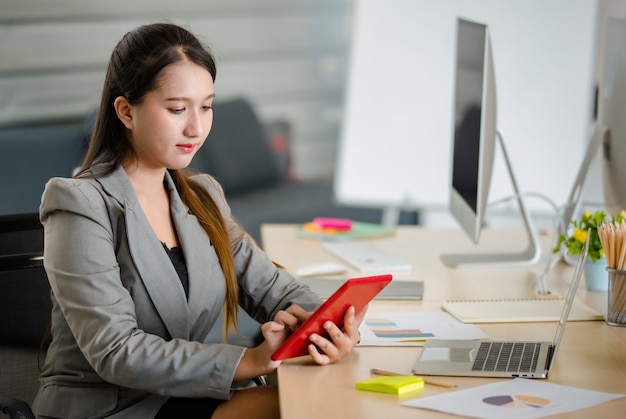 Jonge aantrekkelijke Aziatische vrouw in grijs pak die aan rode tablet werkt in een modern ogend kantoor met een wazige Windows-achtergrond. Concept voor moderne kantoorlevensstijl.