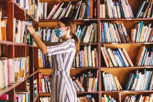 Jonge aantrekkelijke bibliothecaris met gezichtsmasker bij het zoeken naar een boek terwijl hij in de bibliotheek staat tijdens een coronavirus-pandemie.