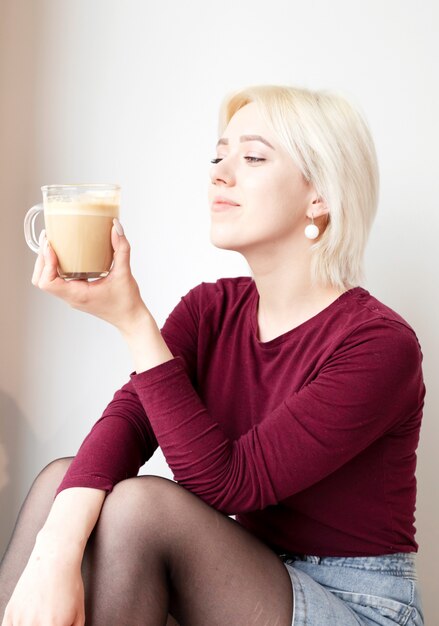 Jonge aantrekkelijke blonde gitl die kop van koffie in ochtend bekijkt.