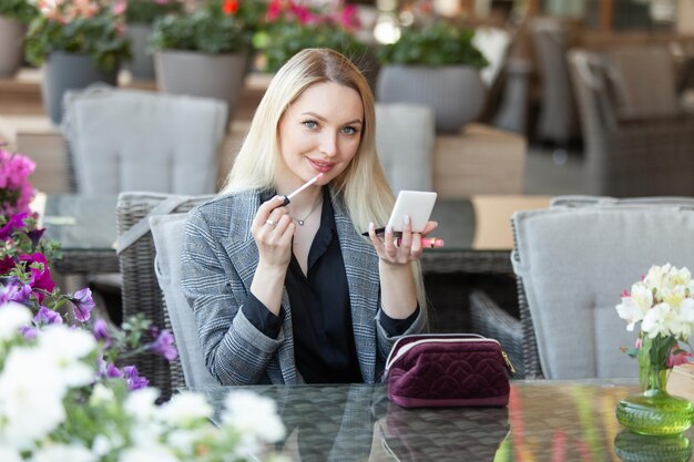 Jonge aantrekkelijke blonde schoonheidsvrouw schildert lippen met glans op het zomerterras van café