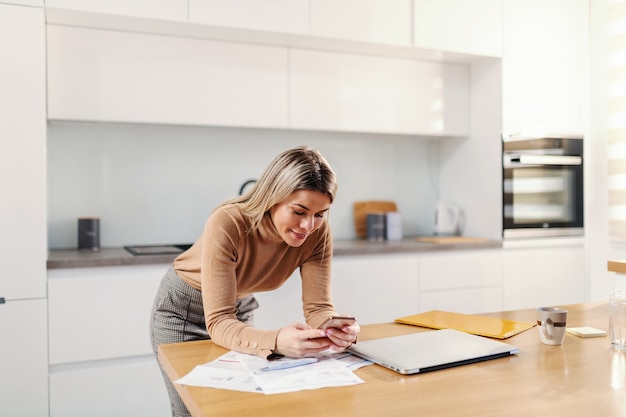 Jonge aantrekkelijke blonde vrouw die op keukentafel leunt en slimme telefoon gebruikt voor e-bankieren.