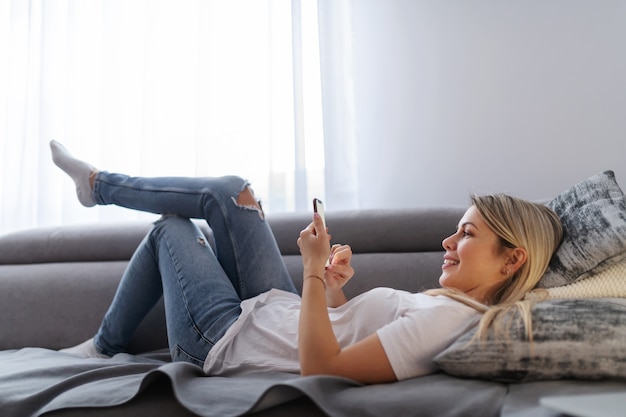 Jonge aantrekkelijke blonde vrouw liggend op de bank in de woonkamer, ontspannen en opknoping op sociale media.