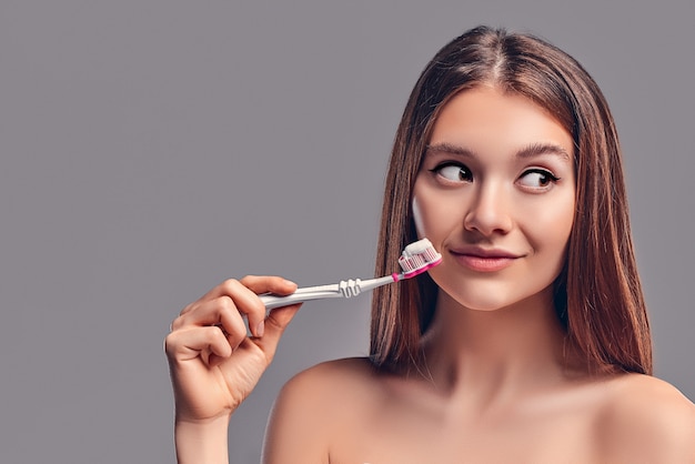 Jonge aantrekkelijke brunette meisje met losse haren maakt gebruik van een tandenborstel en plakken geïsoleerd op een grijze achtergrond.