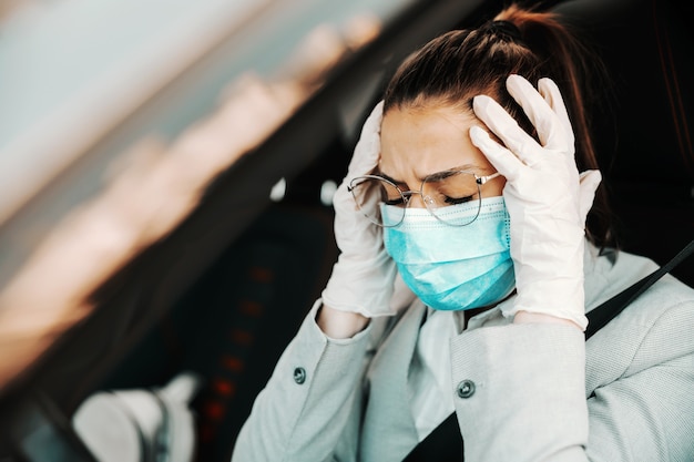 Jonge aantrekkelijke brunette met gezichtsmasker, met rubberen handschoenen om haar hoofd vast te houden omdat ze hoofdpijn heeft terwijl ze in haar auto zit tijdens de uitbraak van het coronavirus.