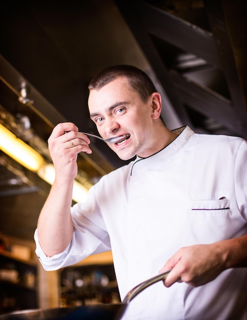 Jonge aantrekkelijke chef-kok die een koekenpan vasthoudt en de gekookte schotel in de keuken van het restaurant test, kijkend naar de camera.