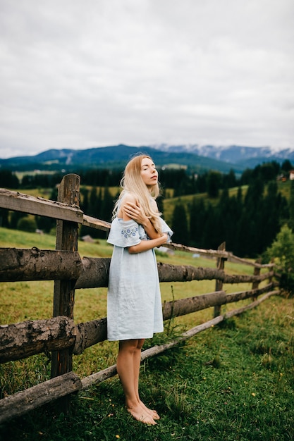 Foto jonge aantrekkelijke elegante blonde meisje in blauwe romantische jurk poseren in de buurt van hek op het platteland