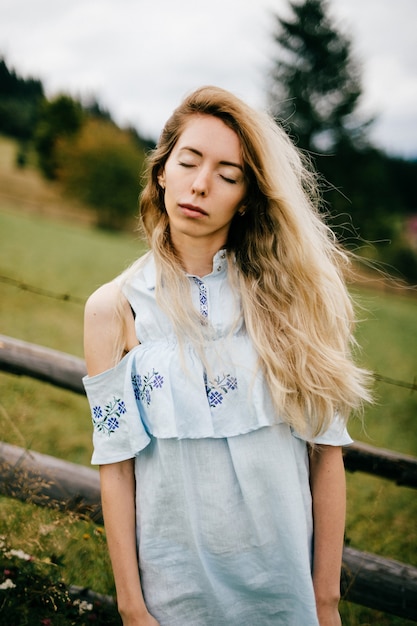 Foto jonge aantrekkelijke elegante blonde meisje in blauwe romantische jurk poseren in de buurt van hek op het platteland