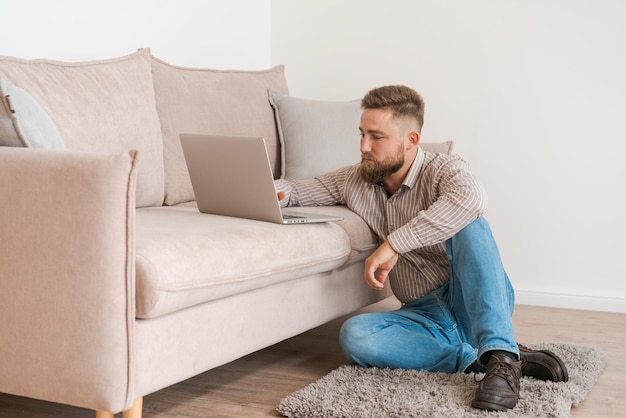 Jonge aantrekkelijke glimlachende bebaarde man die op zijn laptop bladert terwijl hij thuis zit