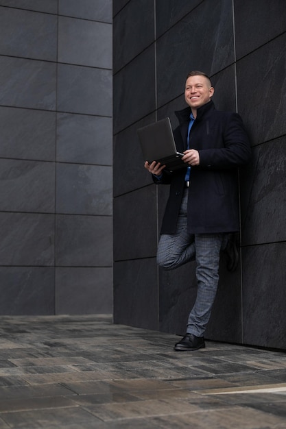 Jonge aantrekkelijke glimlachende zakenman gekleed in zwarte jas staat tegen de stadsbouw met lapt