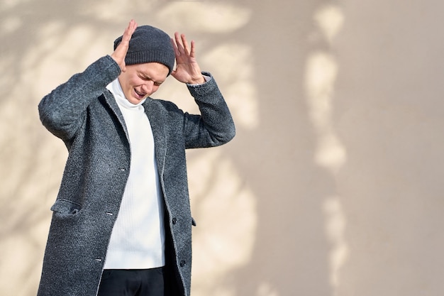 Jonge aantrekkelijke hipster trendy man met gesloten ogen, gekleed in een grijze jas, witte trui en zwarte spijkerbroek hand in hand in de buurt van hoofd en schreeuwen.