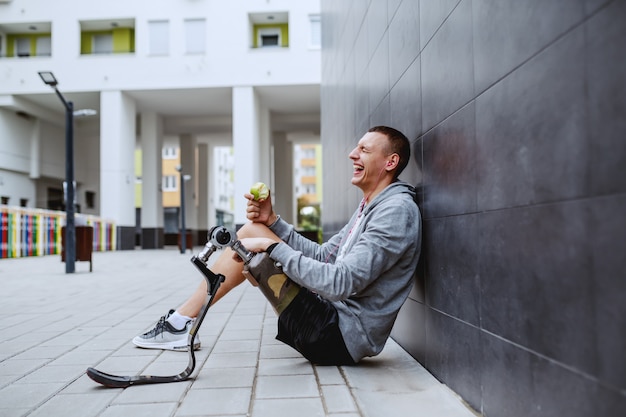 Jonge aantrekkelijke Kaukasische sportman met kunstbeen zittend op de grond, leunend op de muur en het eten van verse appel.