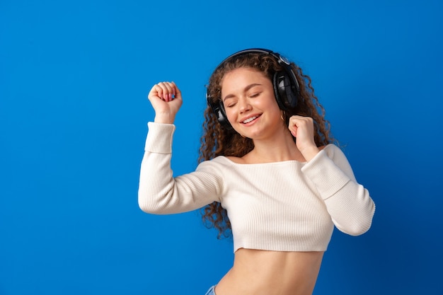 Jonge aantrekkelijke krullende vrouw die naar muziek luistert met een koptelefoon tegen een blauwe achtergrond