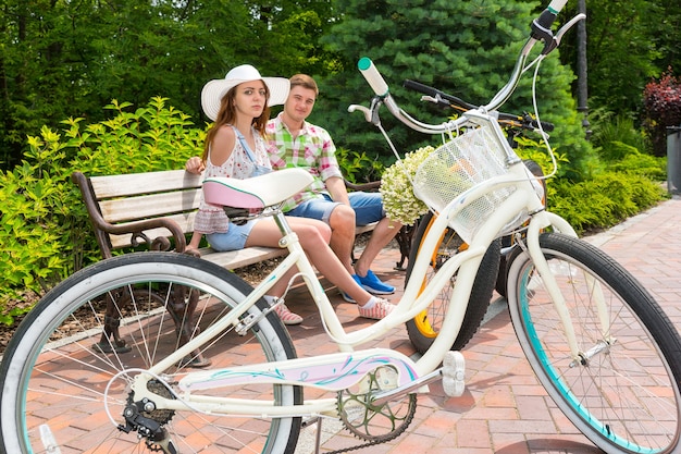 Jonge aantrekkelijke man en vrouw met een witte hoed zittend op een bankje in de buurt van fietsen geparkeerd op bakstenen stoep in een prachtig groen park