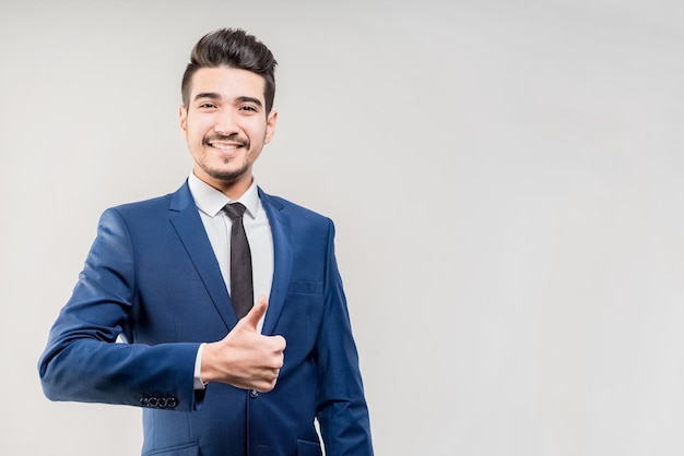 Jonge aantrekkelijke man in een blauw pak duim opdagen op een grijze achtergrond geïsoleerd