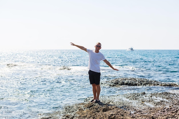 Jonge aantrekkelijke man in zonnebril in een wit t-shirt en korte broek staat aan de kust van de Middellandse Zee