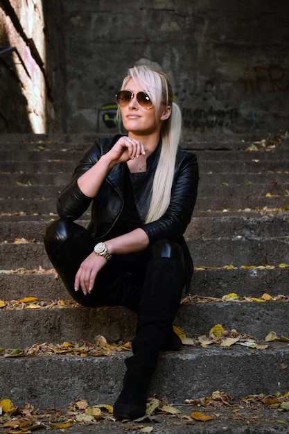 Jonge aantrekkelijke mooie blonde vrouw in zonnebril zittend op stenen trappen en glimlachend