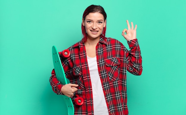 Jonge aantrekkelijke roodharige vrouw die zich gelukkig, ontspannen en tevreden voelt, goedkeuring toont met ok gebaar, glimlachend en houdt een skatebord vast
