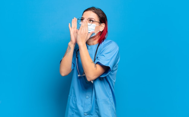 Jonge aantrekkelijke roodharige vrouw die zich gelukkig, opgewonden en positief voelt, een grote schreeuw geeft met de handen naast de mond, roept. ziekenhuis verpleegkundige concept