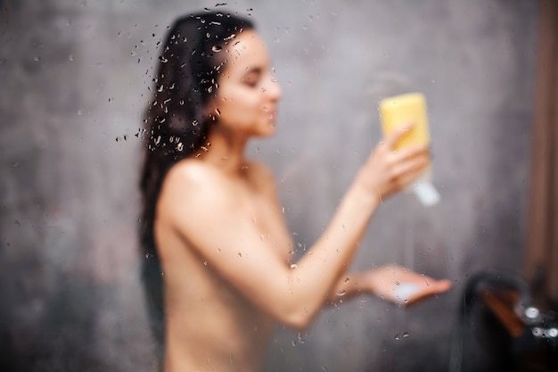 Jonge aantrekkelijke sexy vrouw in douche. Mooi donkerharige model legt wat douchegel bij de hand en glimlacht. Ze staat onder waterstroom. Wazig beeld. Waterdamp op glazen wand.