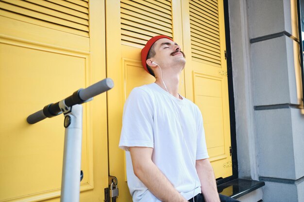 Jonge aantrekkelijke snor man in T-shirt en rode hoed zittend met e-scooter terwijl dromerig muziek luisteren in oortelefoons met gele achtergrond op straat in de stad