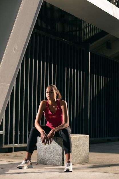 Jonge aantrekkelijke sportvrouw die buiten op de stenen kubus zit en wegkijkt