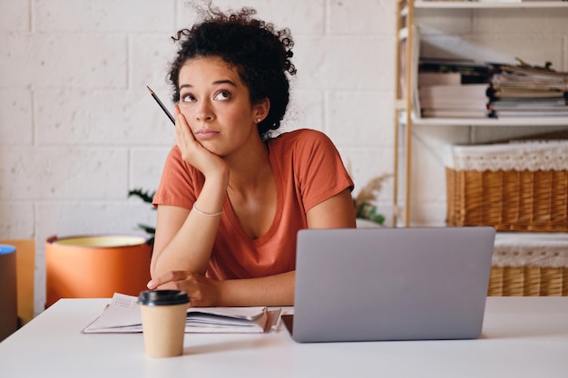 Jonge aantrekkelijke student meisje met donker krullend haar zittend aan de tafel met laptop en kopje koffie om te gaan leunend aan de hand dromerig opzoeken en studeren in modern gezellig huis